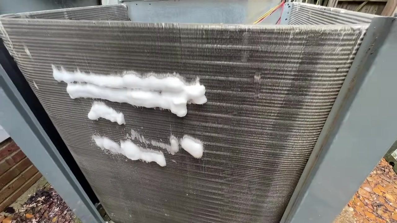 The indoor coil is visible inside the HVAC unit, with foam cleaner already applied in streaks. The HVAC unit cover has been removed, and the setting is outdoors next to a building.