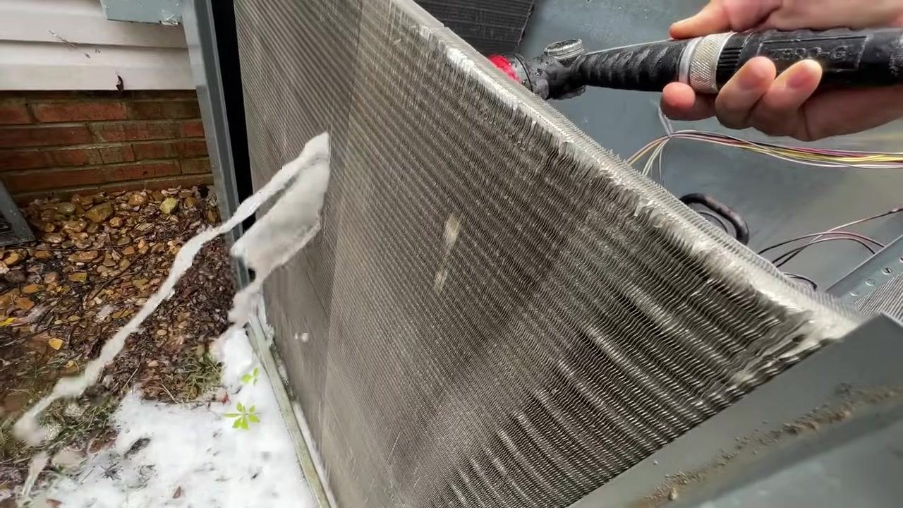A person uses a hose with a spray nozzle to rinse foam and debris from the coil, with foam running off into the ground below.