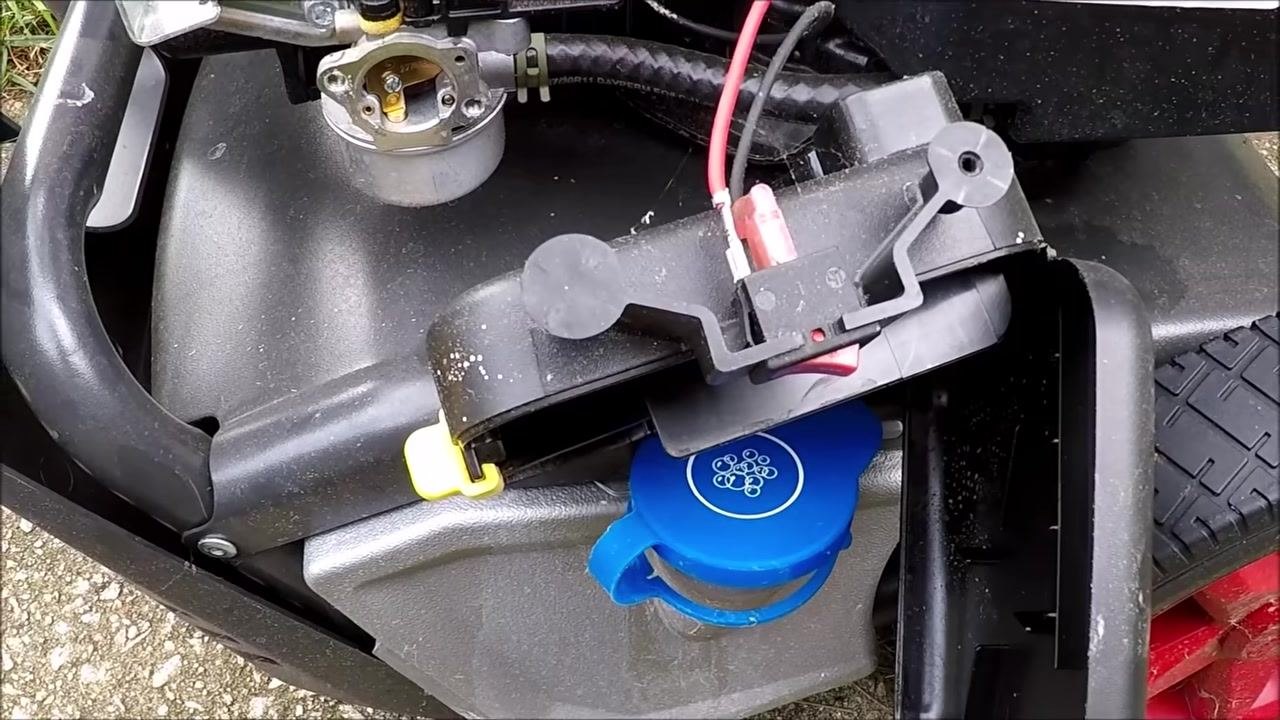 Close-up of the removed air filter door assembly resting on the pressure washer frame, showing the blue detergent cap, yellow clip, attached red and black wires, and the exposed carburetor.