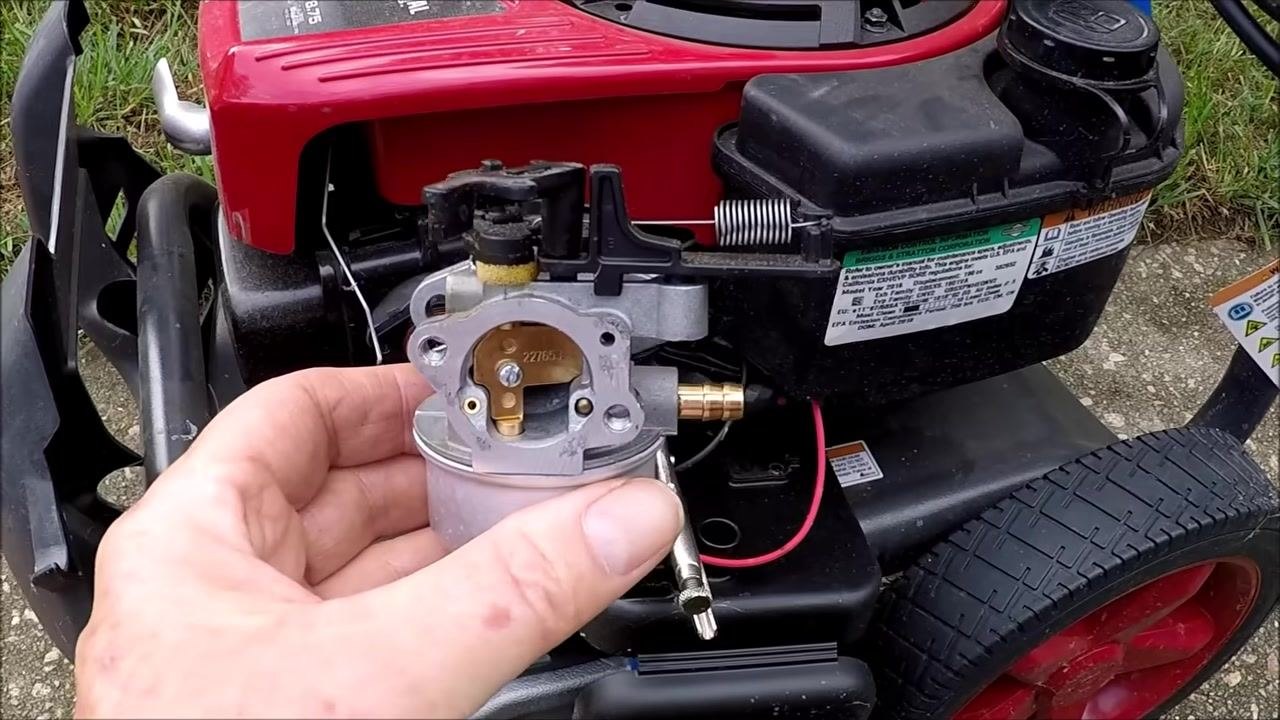 Carburetor held over a white plastic container with yellowish fluid visible, float bowl removed, hands holding both parts.