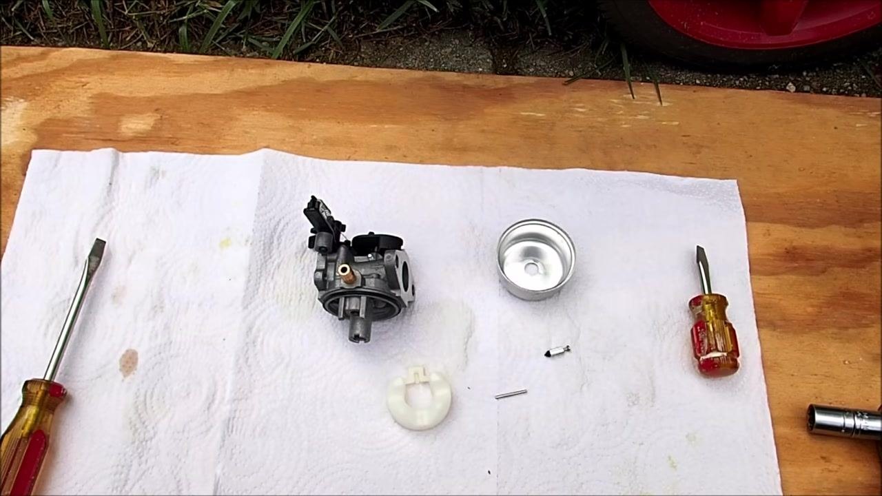 Carburetor body, float bowl, float, float pin, needle, two screwdrivers, and a socket laid out on a white towel on a wooden board outdoors.