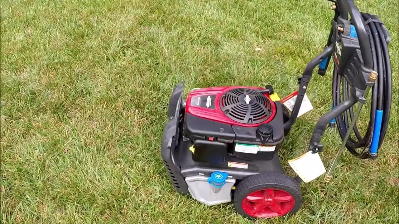 Angled top view of the power washer on grass with all components visible and the engine running, hose and handle attached.