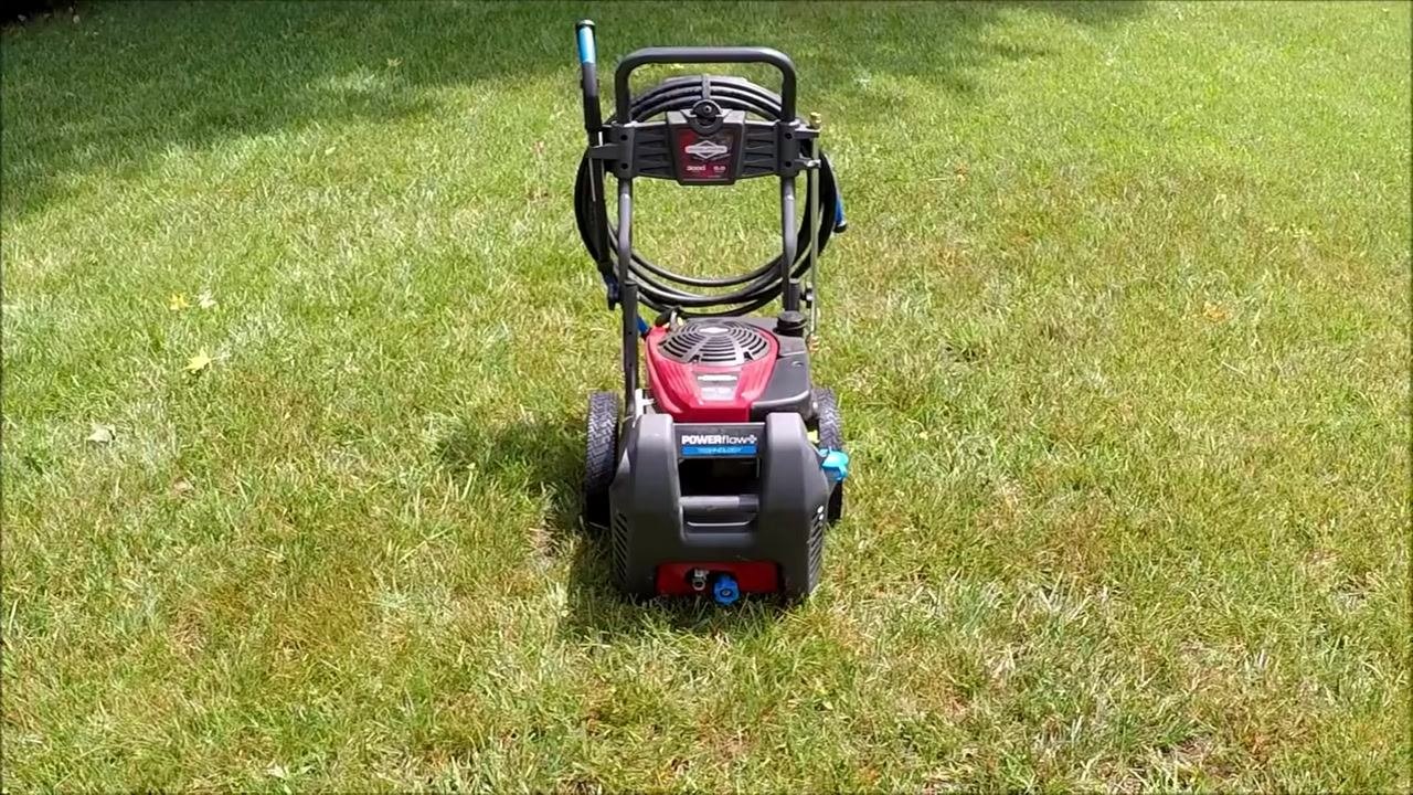 Side view of the pressure washer showing the engine, exhaust, hose reel, and red wheels on grass, with tags attached and the spray gun mounted.