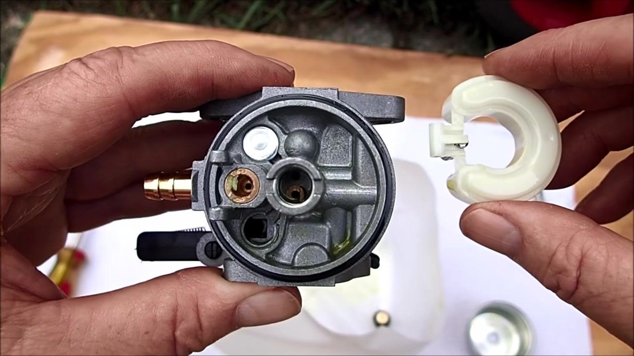 Hands holding the carburetor with the float removed, showing a close-up of the main jet and fuel passages, with a screwdriver and float bowl visible in the background.