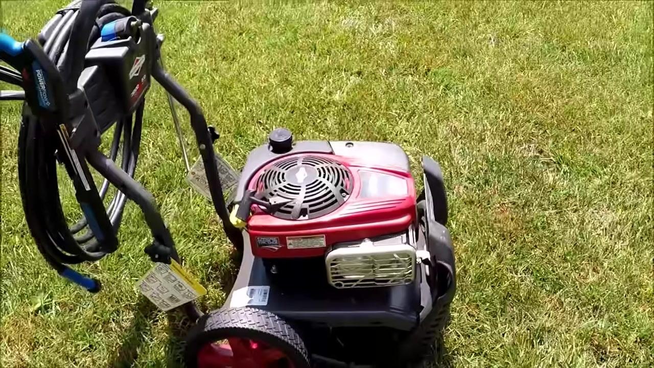 Pressure washer on grass, showing large rear wheels, hose reel, spray gun, and engine cover with visible tags and labels.