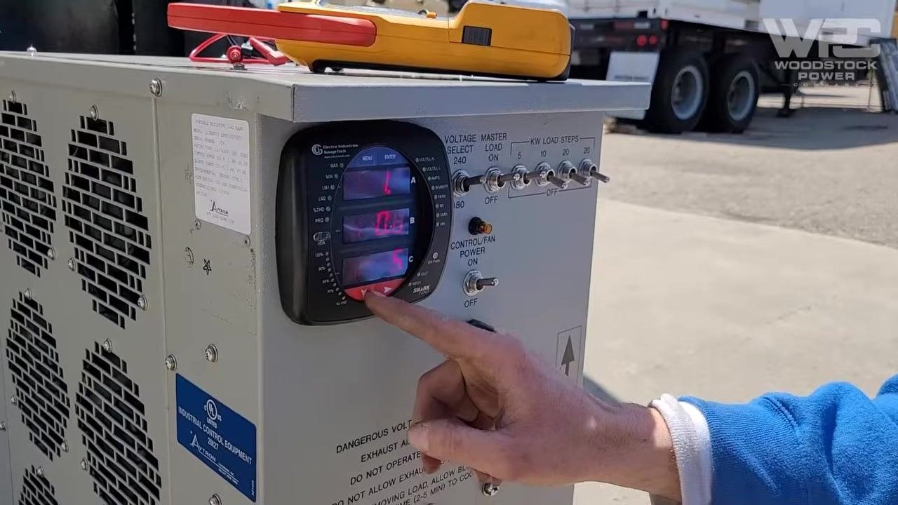 Technician pointing at the digital ammeter on the load bank panel, showing current readings for each phase with load step switches and warning labels visible