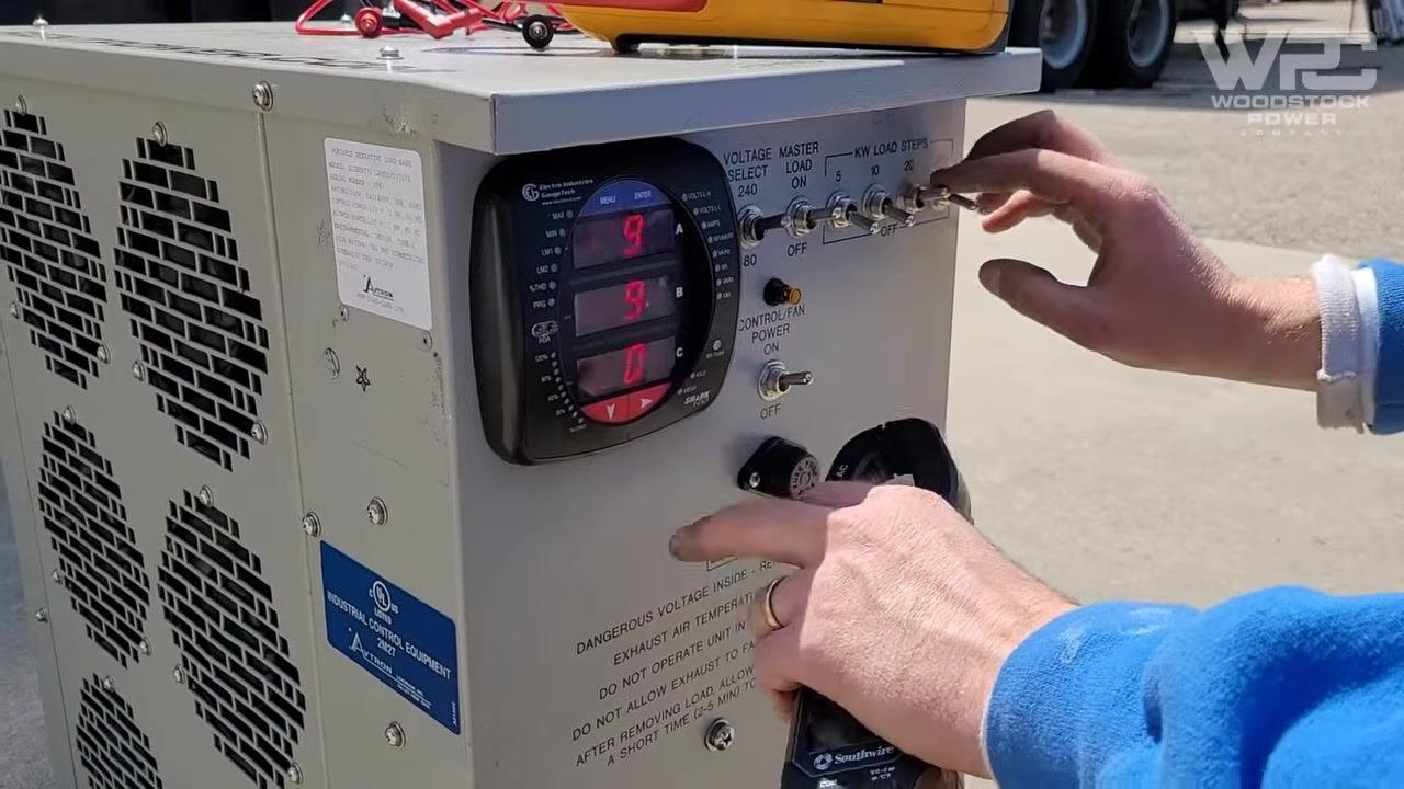 Load bank control panel with digital display, toggle switches for voltage and load steps, warning labels, and a hand adjusting the controls