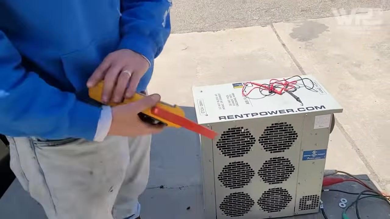 Technician holding a clamp meter and test leads next to the load bank, preparing for measurement
