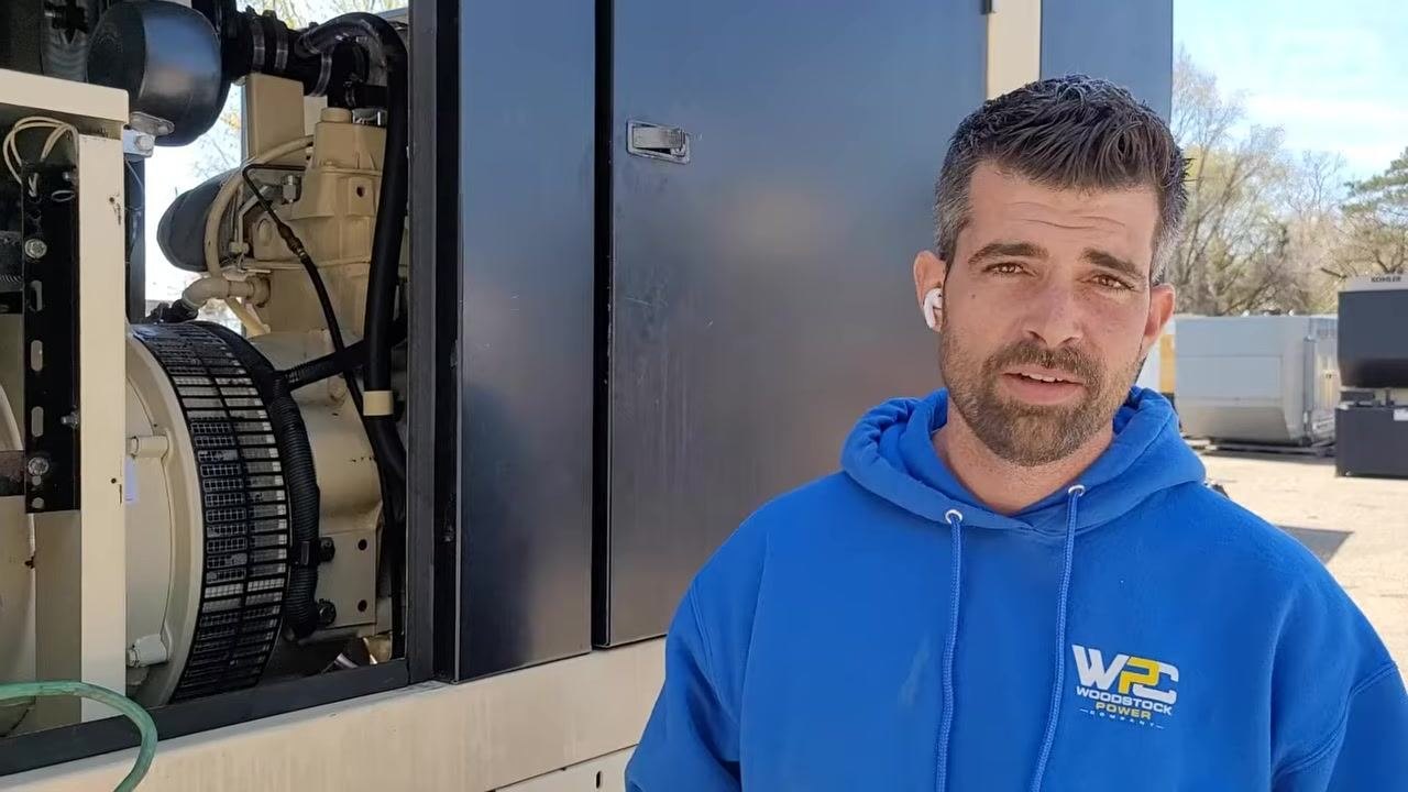 Person in blue Woodstock Power hoodie standing next to generator, speaking to camera