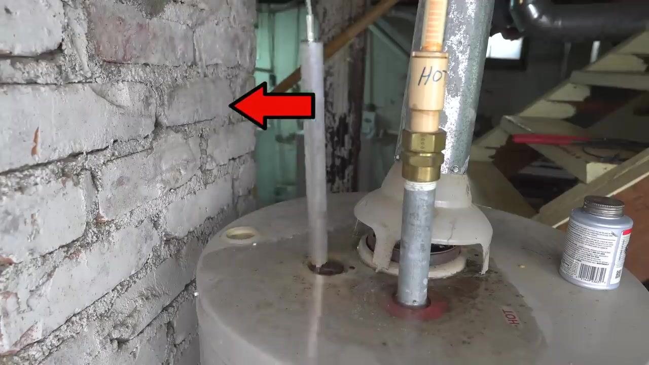 A person using a yellow impact wrench to remove the anode rod from the top of a water heater, next to a pipe labeled "HOT" in a basement setting.
