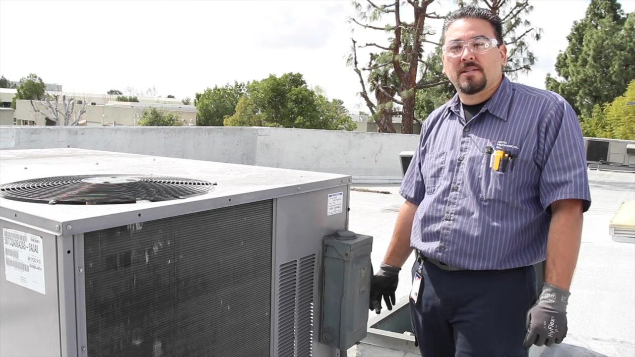 Technician standing beside the HVAC unit, addressing the camera, wearing safety glasses and gloves, with company branding visible on the shirt.