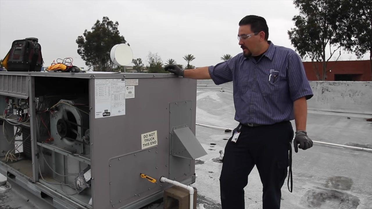 Technician standing on a rooftop next to an open HVAC unit, pointing at the unit while discussing economizer functionality, with internal components and specification labels visible.