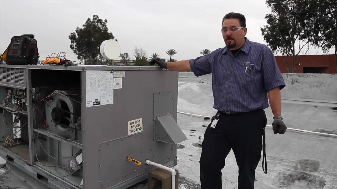 Technician standing next to an open rooftop HVAC unit, with tools and warning labels visible.
