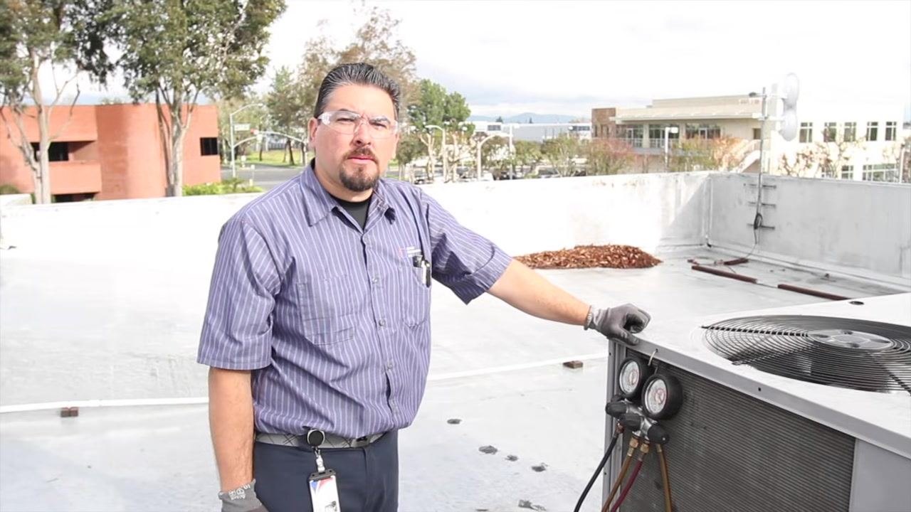 Technician standing next to an energized rooftop HVAC unit, with pressure gauges attached and safety gear on.