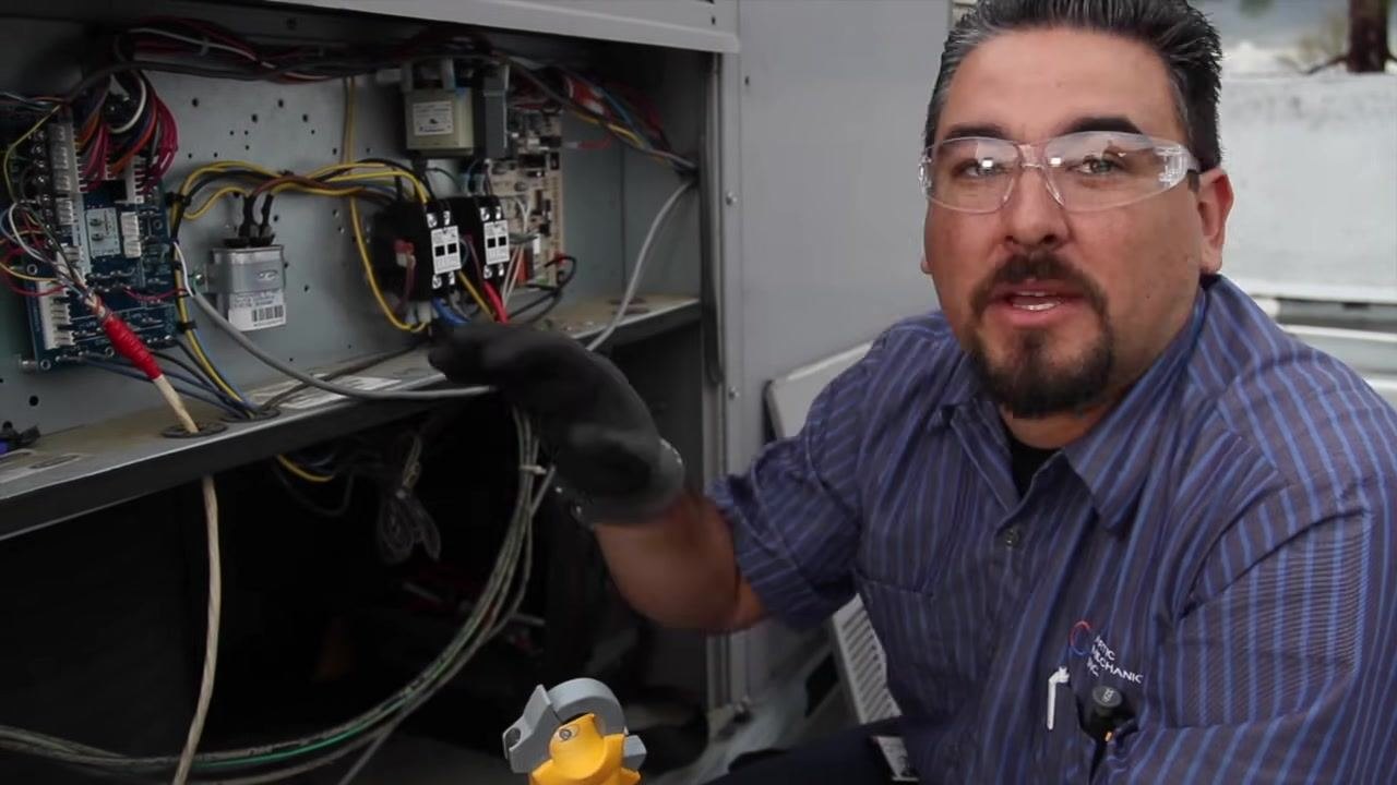 Technician explaining the need to replace the capacitor, holding a clamp meter inside the open electrical compartment.