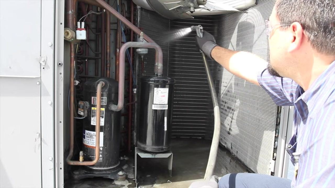 Technician spraying water directly onto the coil inside the HVAC unit, with internal copper piping and the compressor visible.