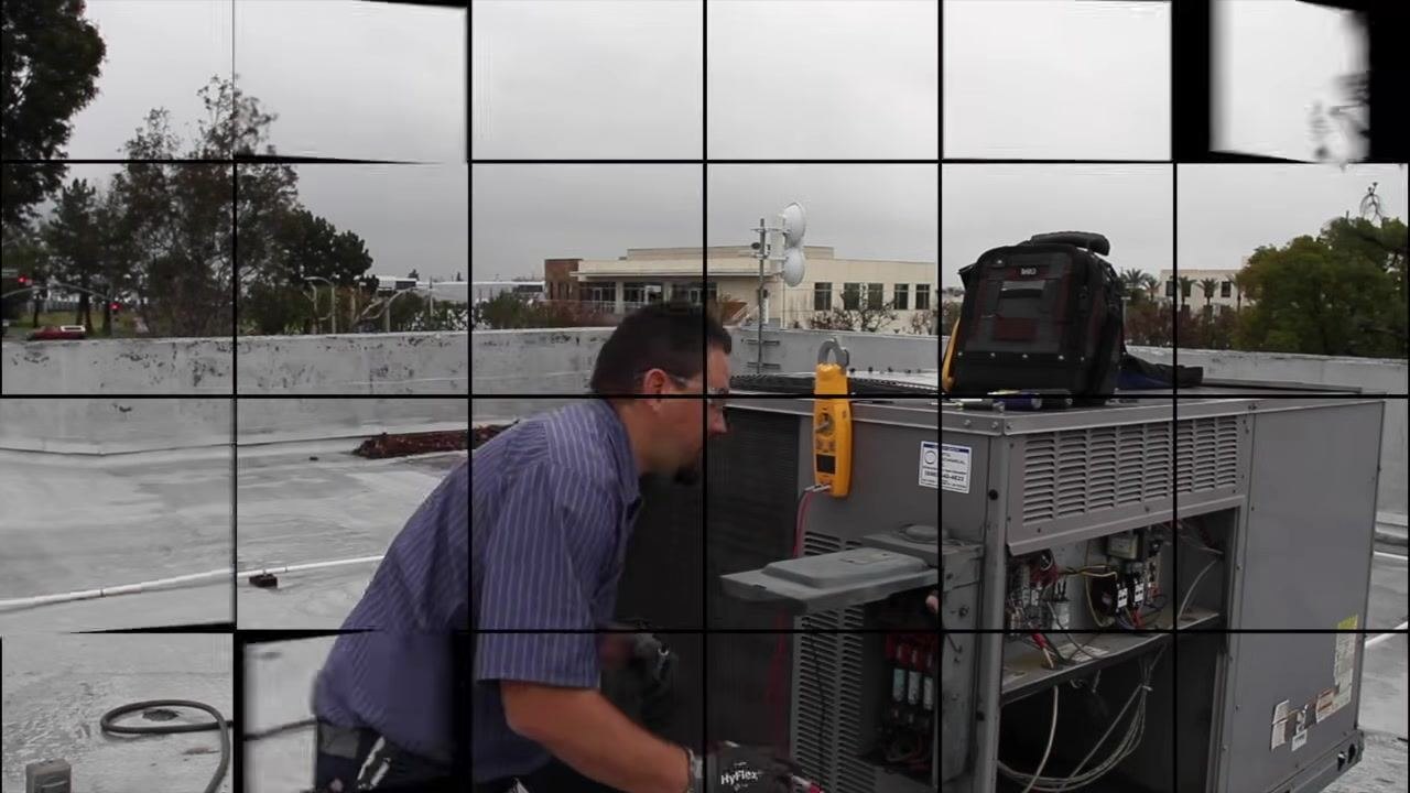 Technician standing next to an HVAC unit on a rooftop with a multimeter and tool bag on the unit; the access panel is open, exposing internal components.