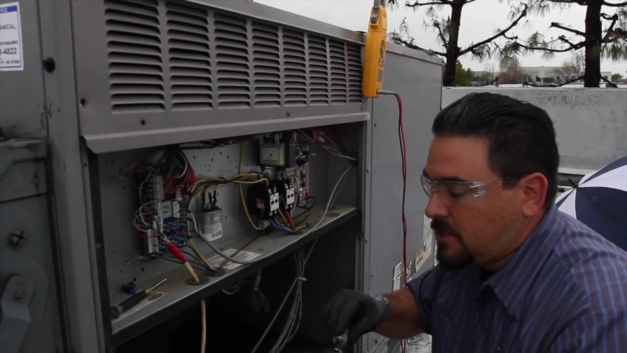 Technician inspecting and tightening wire lugs on contactors inside the HVAC unit's electrical compartment, with a multimeter clipped to the unit.