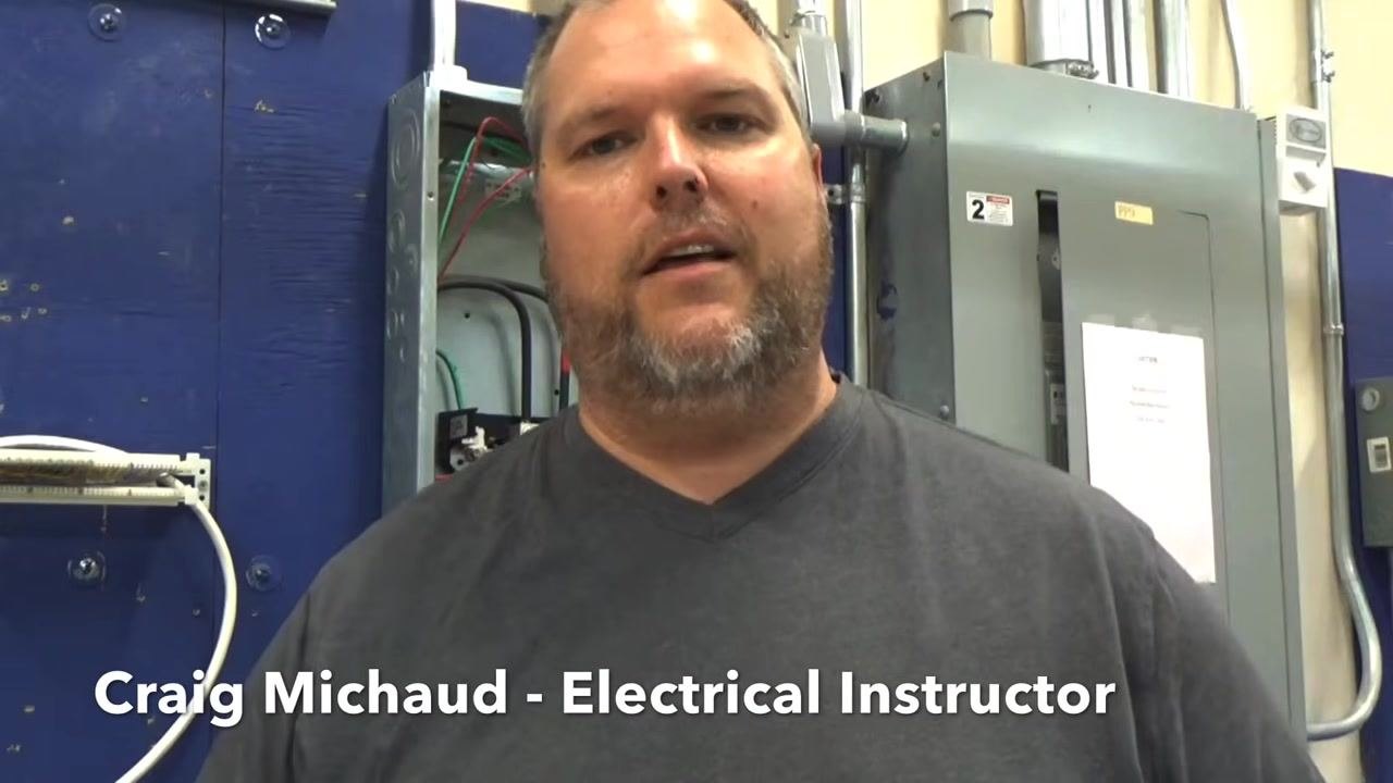 Instructor holding a GFCI/Arc Fault combo breaker, showing the device label and test button, with electrical panels and conduit visible in the background.