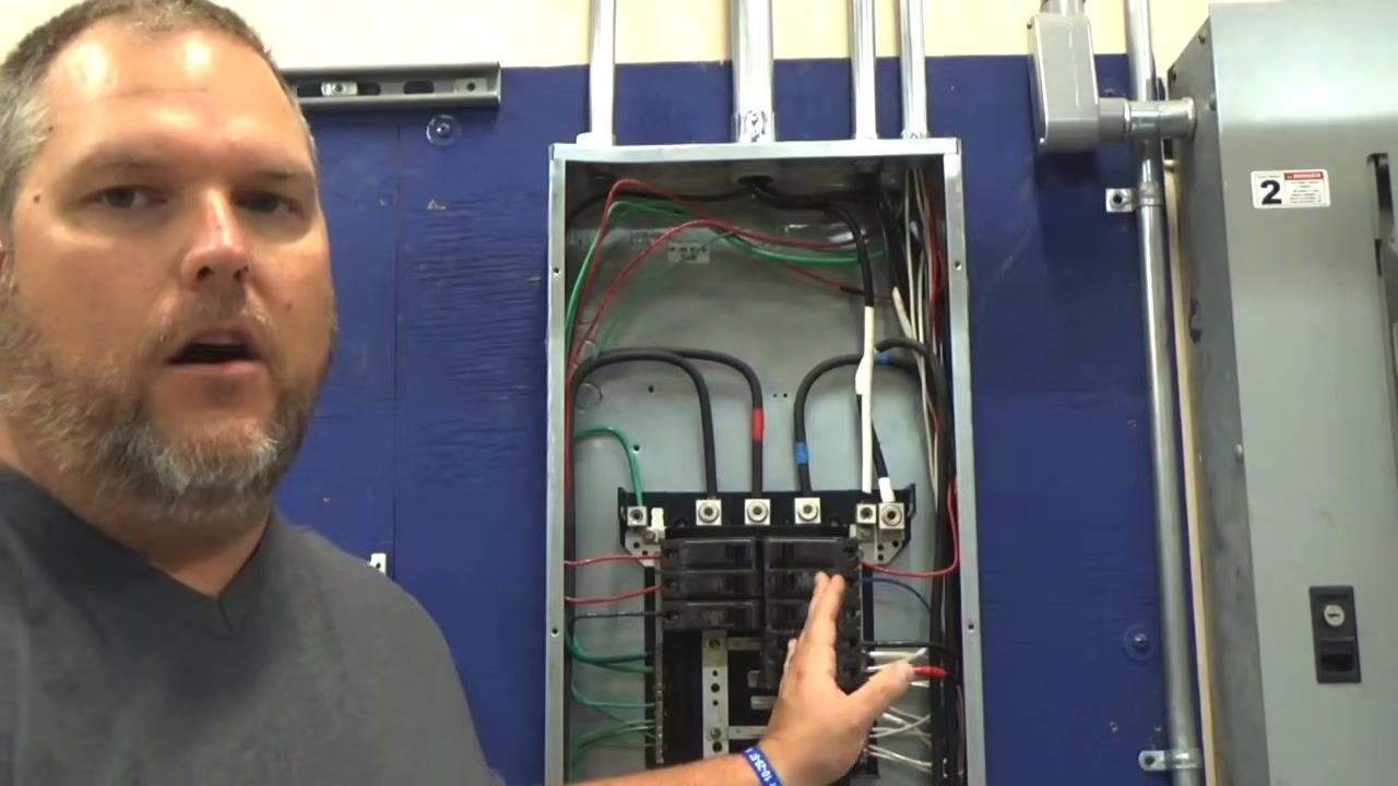 Instructor gesturing toward the open electrical panel, indicating the slot where the GFCI breaker will be installed, with the neutral bar and wiring visible.
