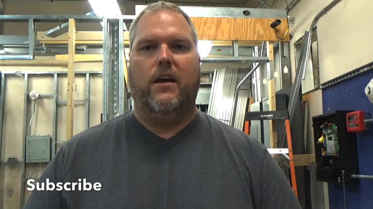 Instructor standing in a workshop, facing the camera, with exposed framing, electrical panels, a step ladder, and the word "Subscribe" in the lower left corner.