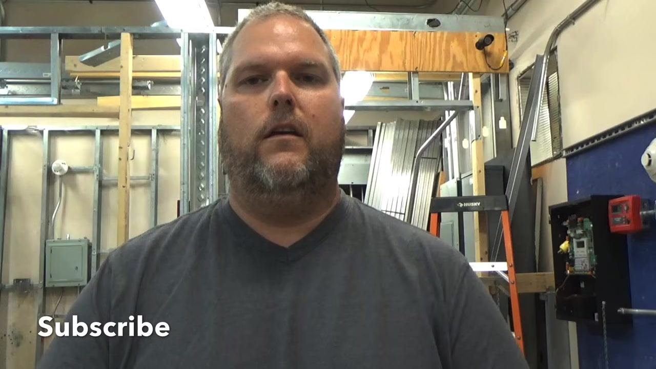 Instructor standing in a workshop, facing the camera, with electrical panels and tools in the background.