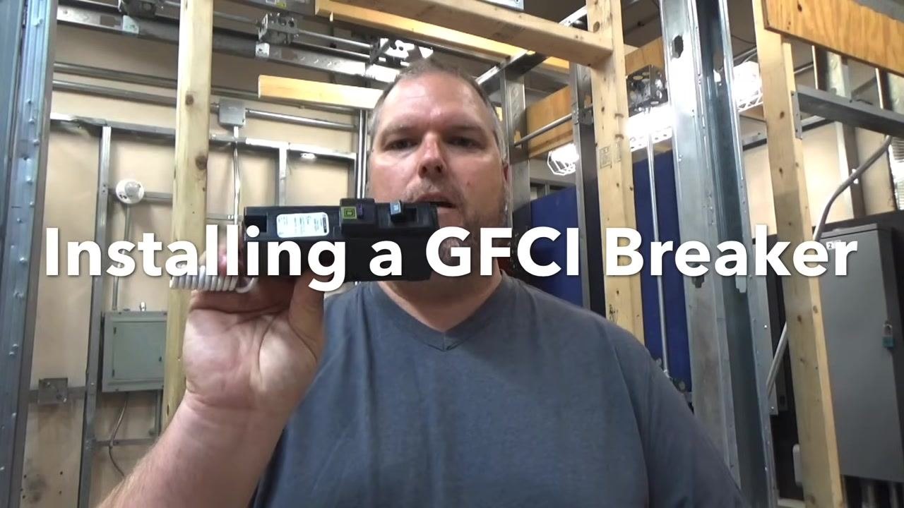 Instructor holding a GFCI breaker, with the text 'Installing a GFCI Breaker' overlaid. Electrical panels and conduit are visible in the background.