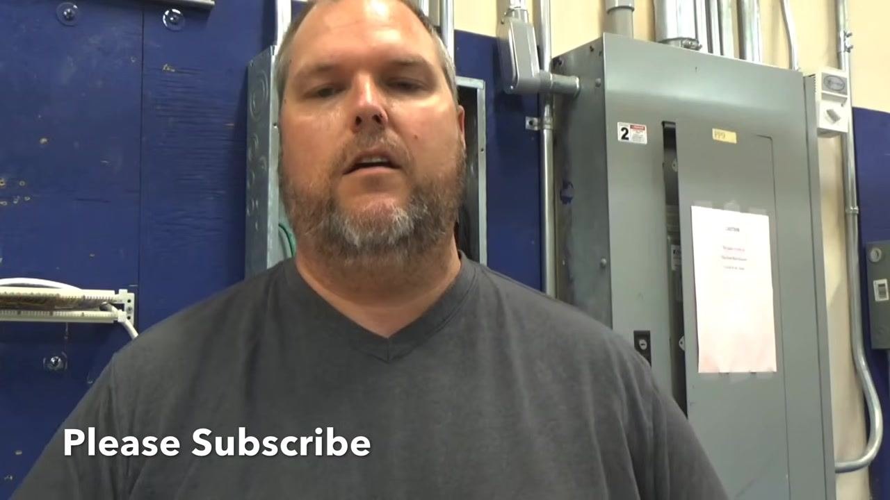 Instructor standing next to an open electrical panel, discussing skill level and safety, with the panel interior, breakers, and wiring visible.