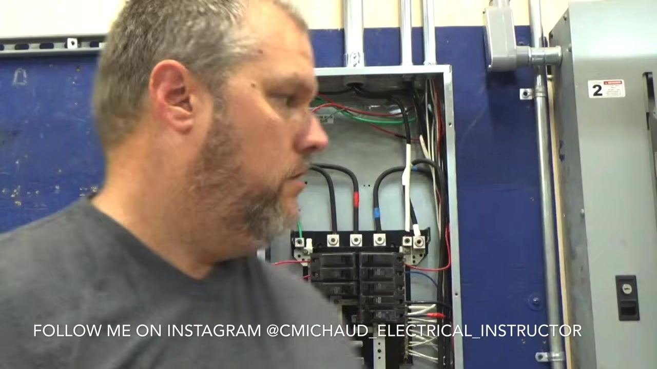 Instructor holding a GFCI breaker close to the camera, pointing out the trip button and internal mechanism, with the electrical panel visible in the background.