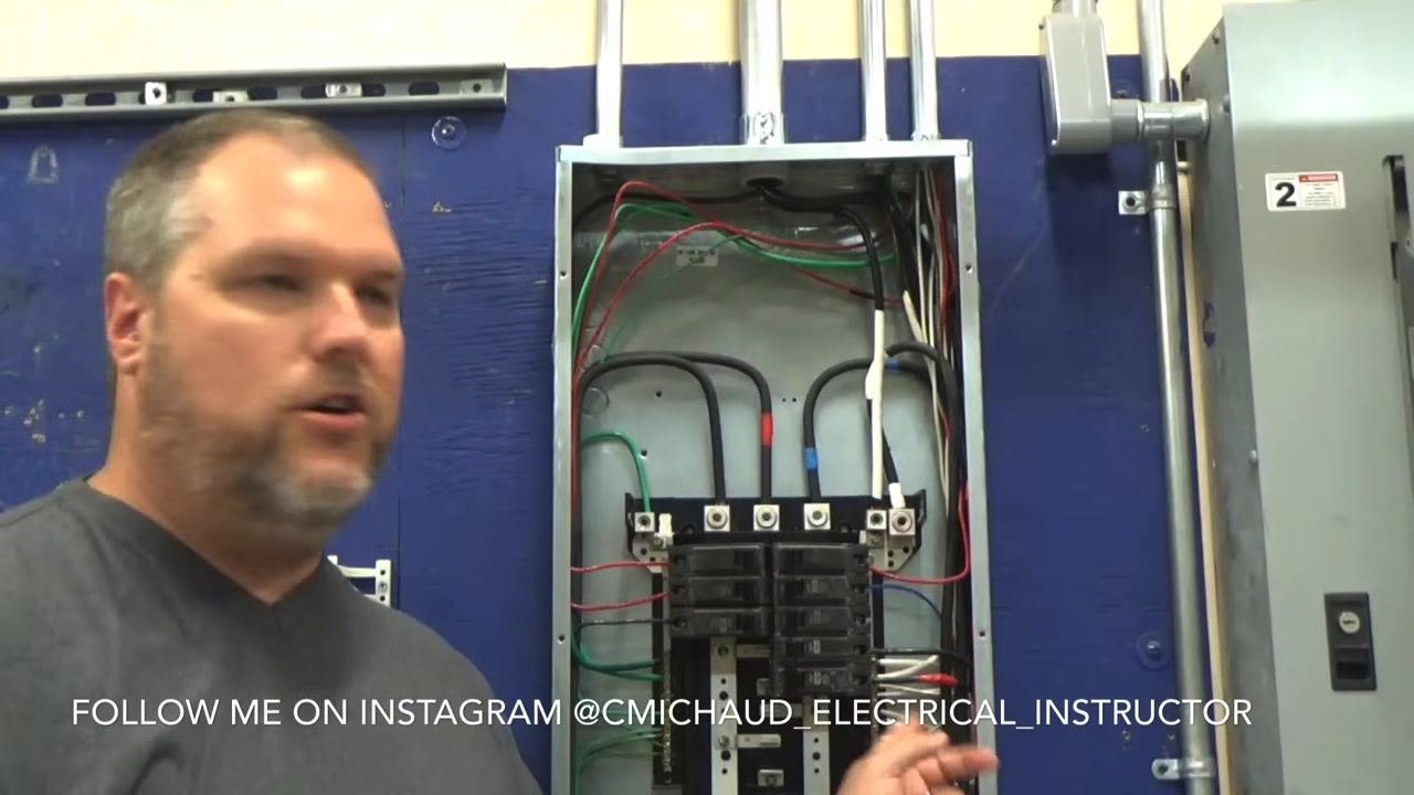 Instructor gesturing toward the open electrical panel, explaining the difference between standard and GFCI breaker wiring.
