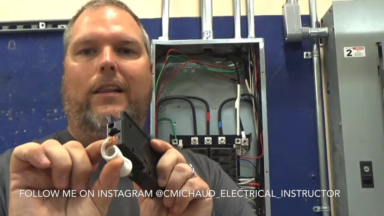 Instructor holding a GFCI breaker, clearly showing the white coiled pigtail wire that will be landed on the neutral bar.