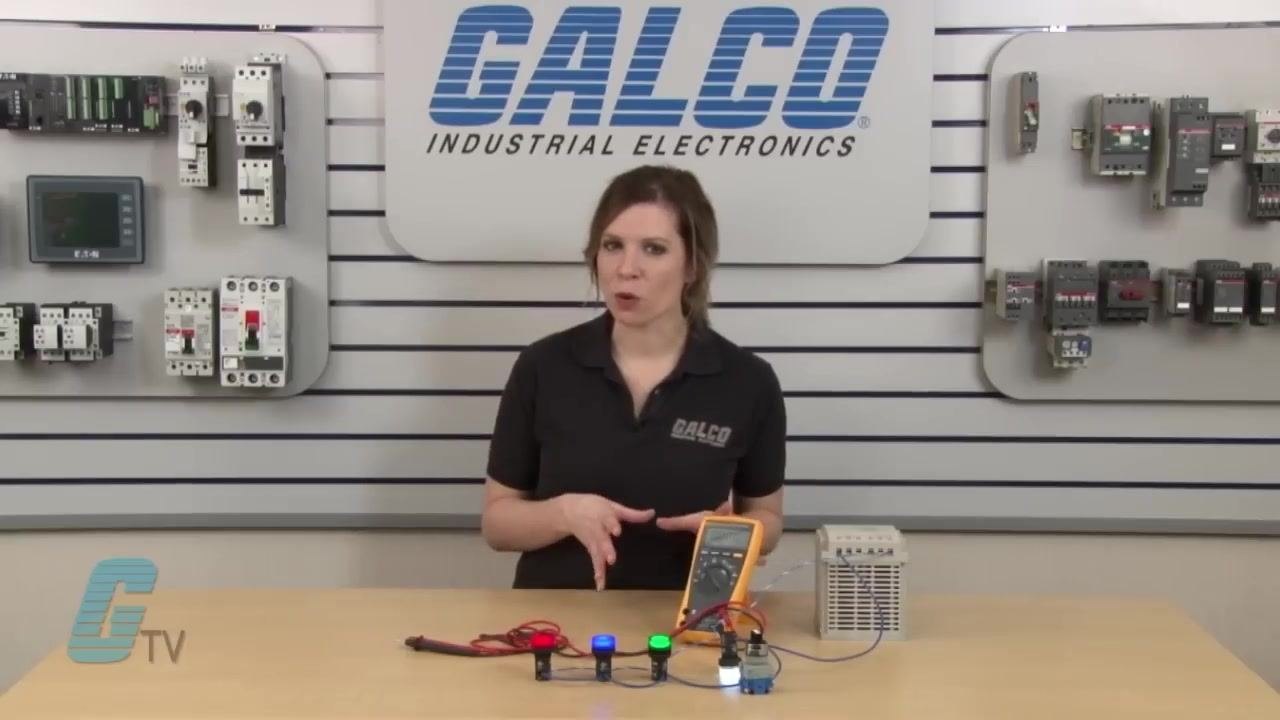 A hand pointing to the demonstration setup, highlighting the multimeter and pilot lights before the voltage measurement.