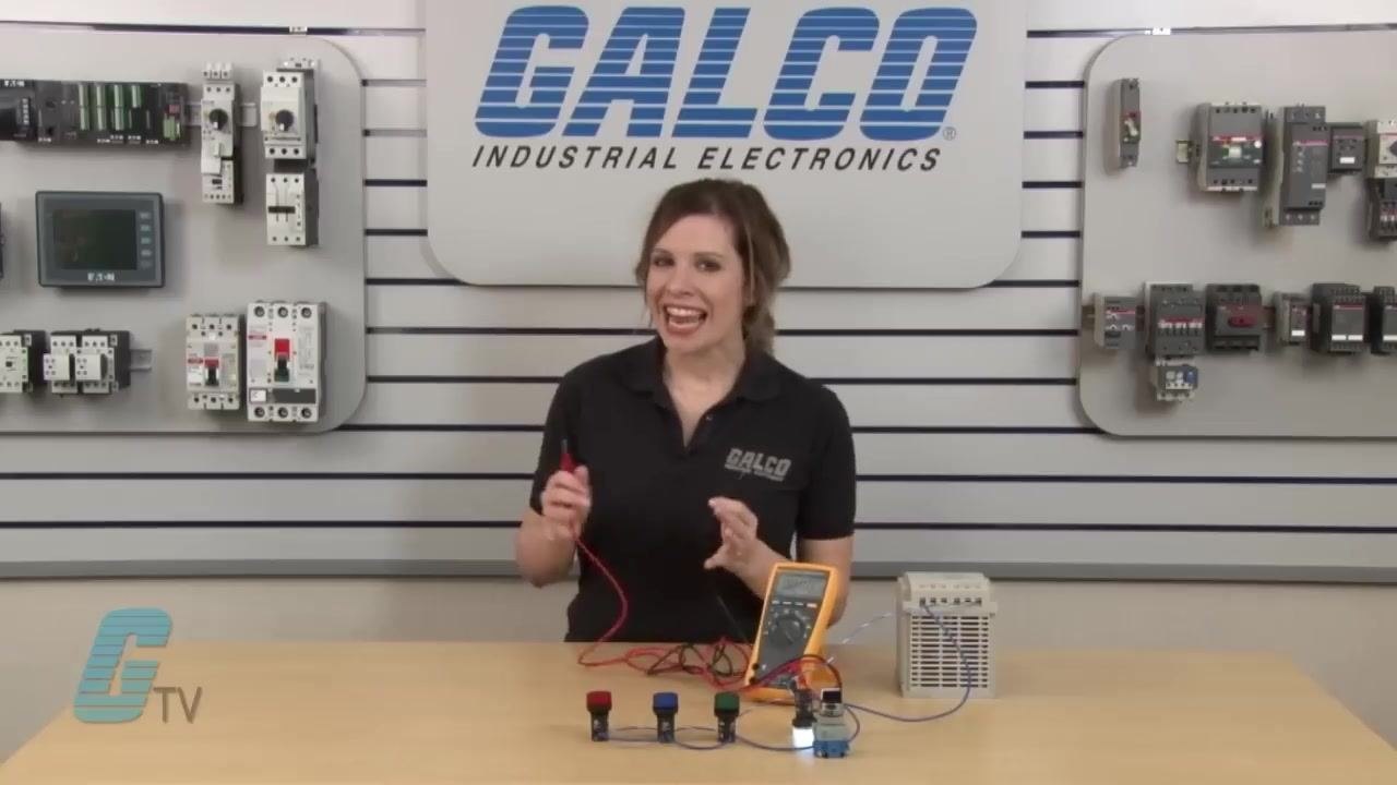 Multimeter leads held up in preparation for the current measurement, with the pilot lights, multimeter, and power supply visible on the demonstration table.
