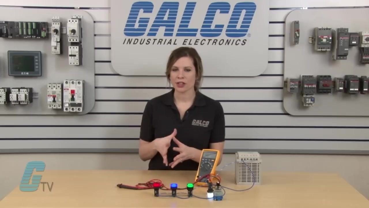 Multimeter probes touching the terminals of the red and blue pilot lights, with the multimeter display active and other pilot lights visible in the background.