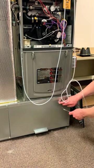 A technician in a blue shirt stands next to an open HVAC furnace. The furnace has visible wiring, a digital dual port manometer attached, and tubing ready for measurement, with the technician preparing to begin the measurement process.