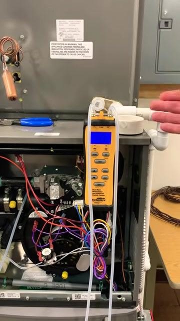 Technician gesturing toward the furnace and manometer, emphasizing the importance of referencing the blower curve chart, with the furnace open and the manometer actively displaying readings.