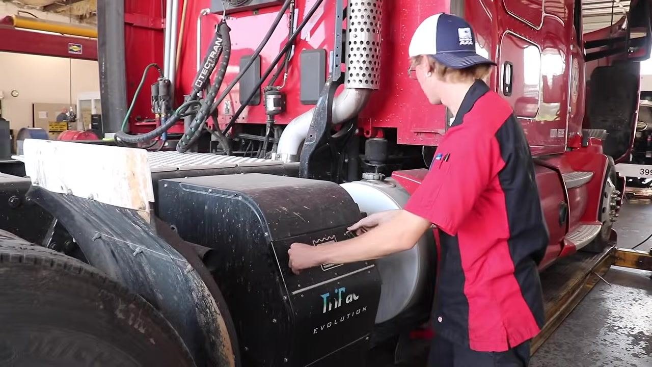 Technician inspecting the side compartment of a red semi-truck, preparing to check tires, airbags, and brakes, with the fuel tank and "TriPac Evolution" side fairing visible.