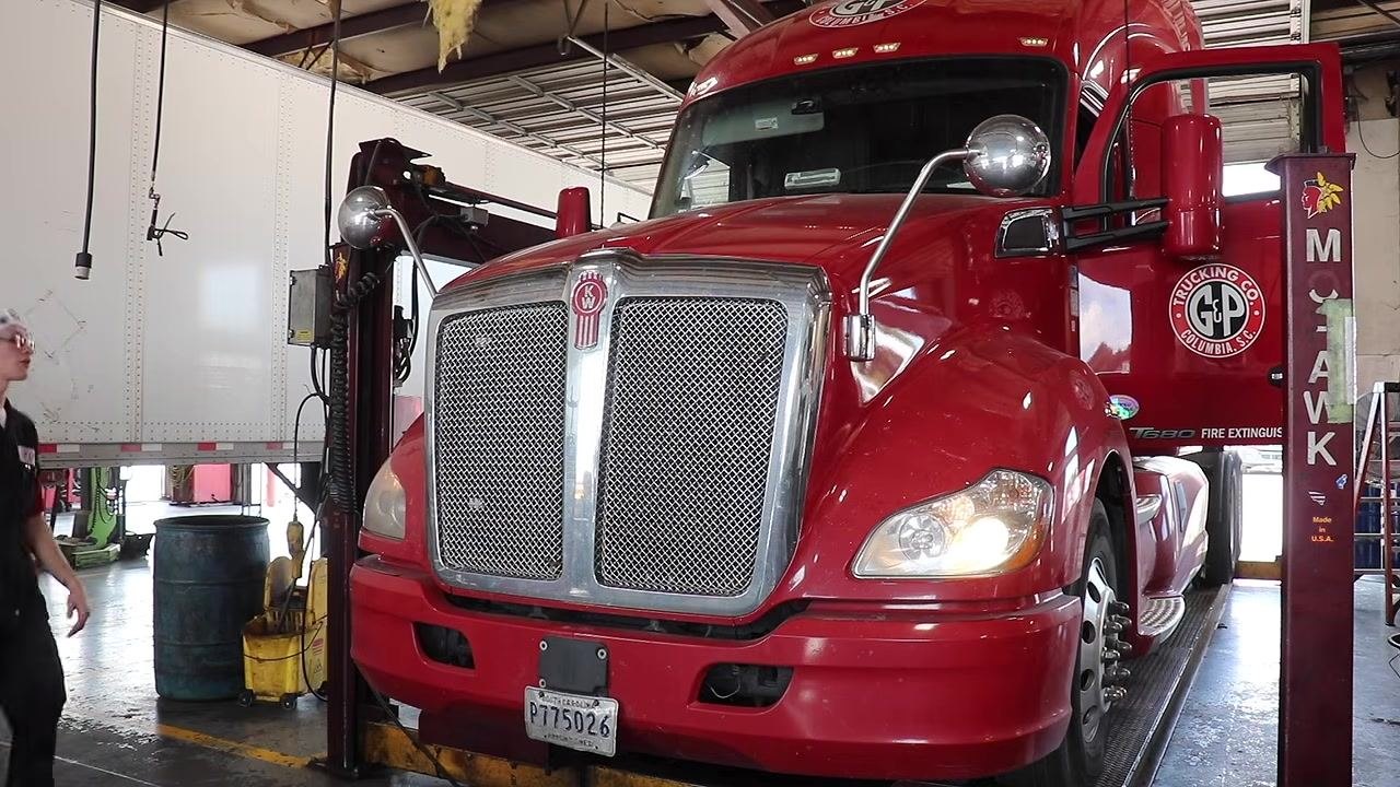 Technician holding a trailer light tester and explaining its use, with other trucks and equipment visible in the shop.