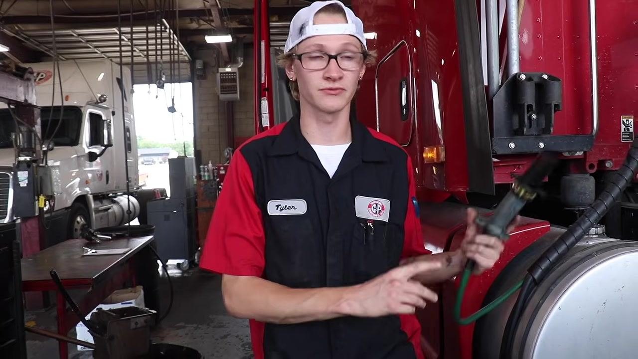 Technician holding a trailer light tester and pointing to the connector, standing beside a red semi-truck in the maintenance shop.