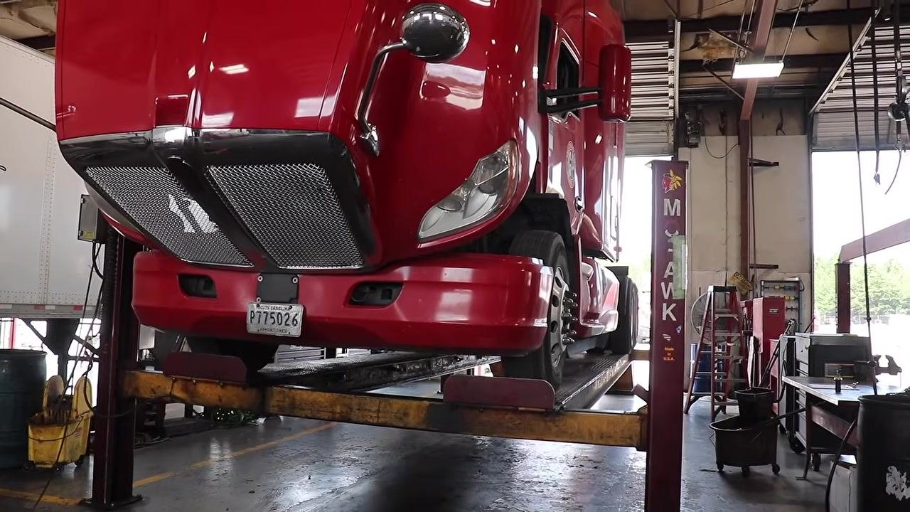 Technician under the truck, shining a flashlight and inspecting engine components, pointing at wiring and hoses.