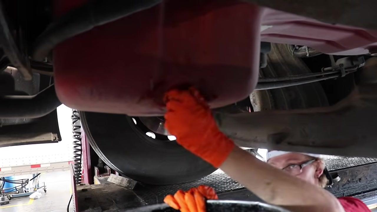 Technician under the truck, pointing at the oil drain plug on the oil pan, preparing for the oil change.