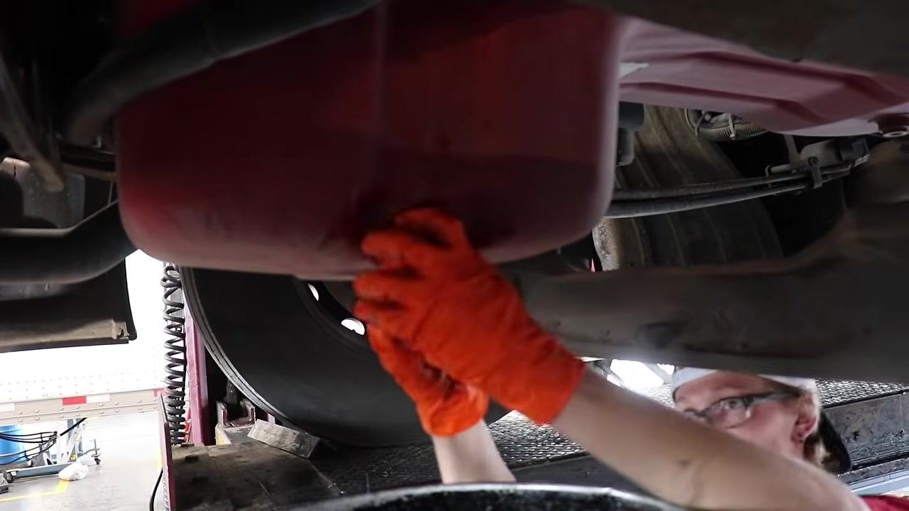 Technician wearing orange gloves, unscrewing the oil drain plug from the bottom of the oil pan.