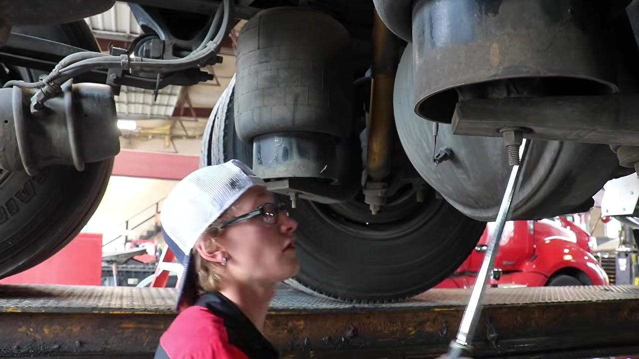 Technician at the rear wheel, using a ratchet to remove the hub oil plug, with suspension and brake components visible.