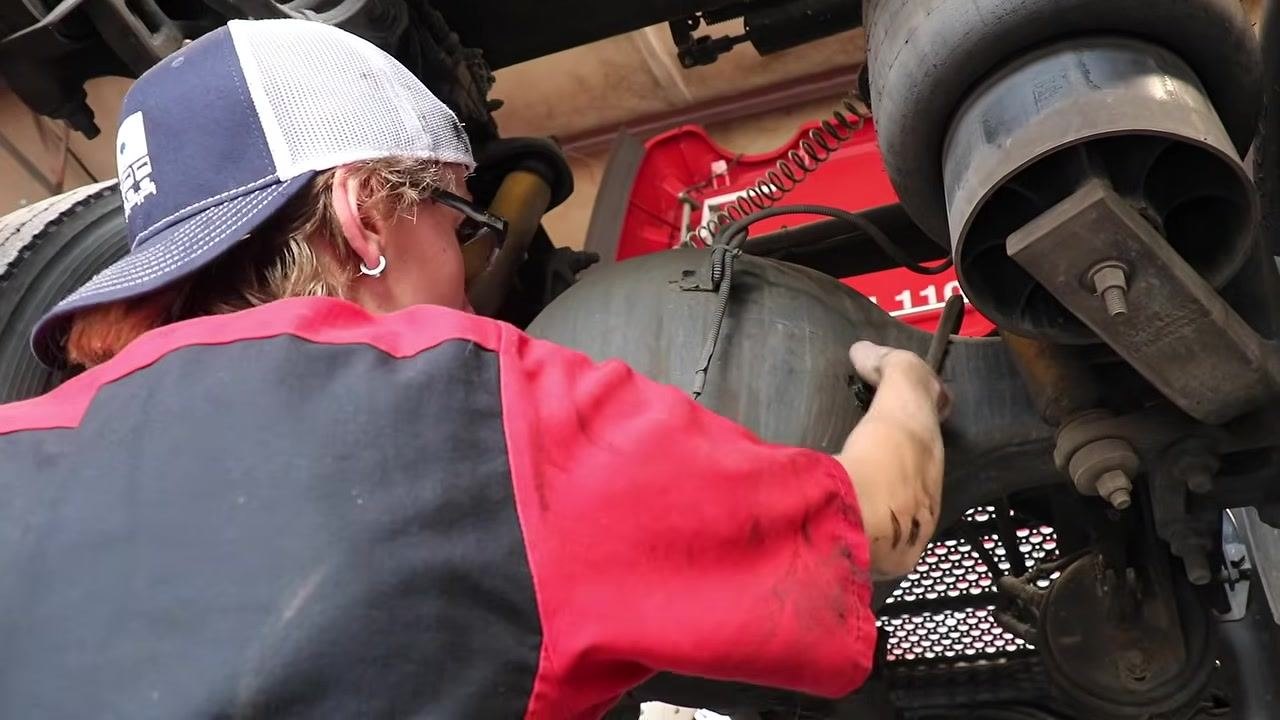 Technician using a wrench to remove a fill plug from a large axle housing under the truck, with suspension and brake components visible.