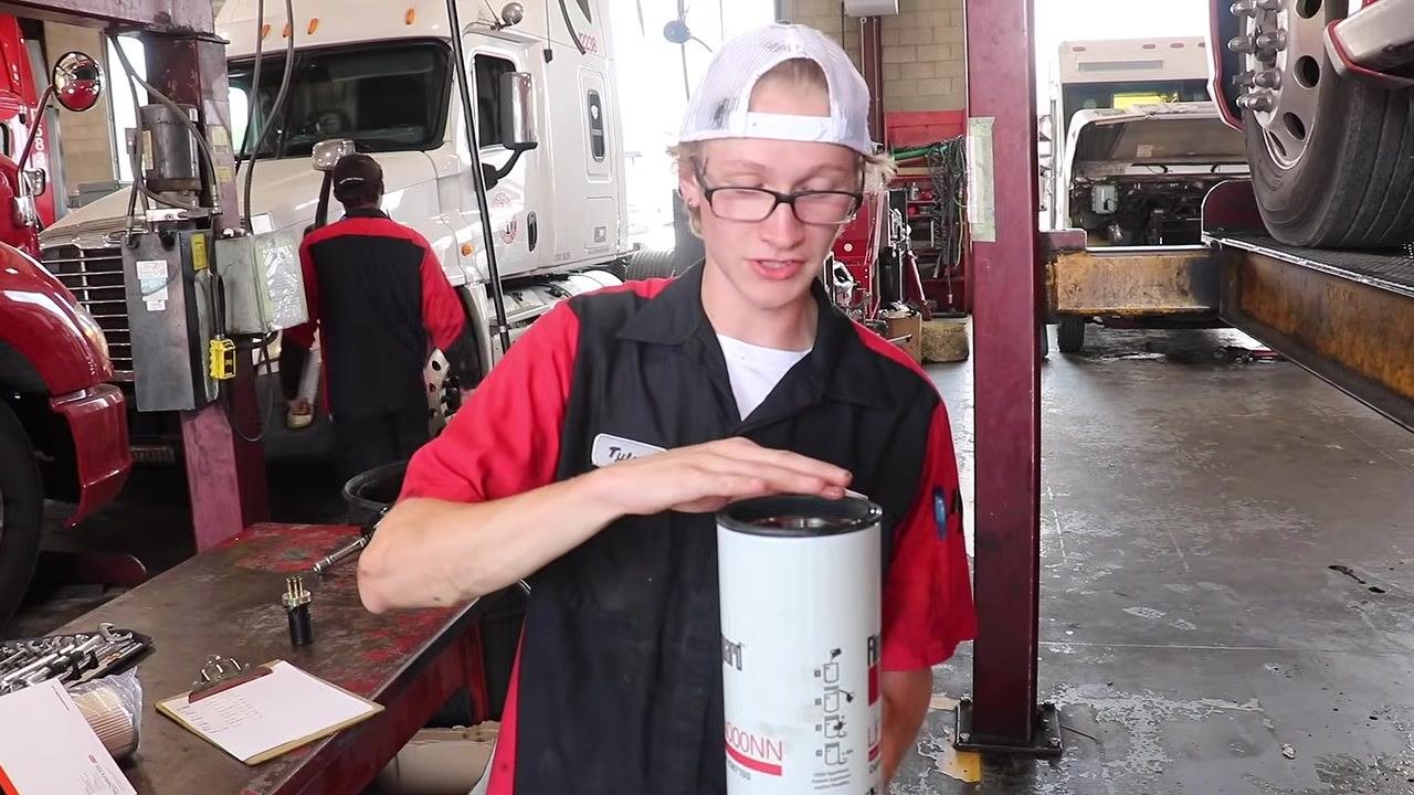 Technician applying oil to the gasket of a new oil filter with his finger, with the filter upright on a workbench.