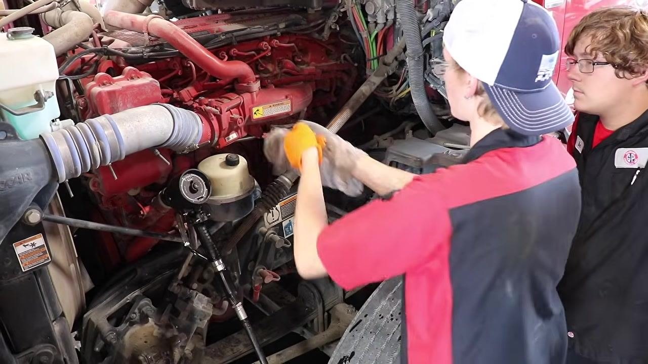 Technician checking the oil level with a dipstick and wiping it clean, with the engine compartment visible.