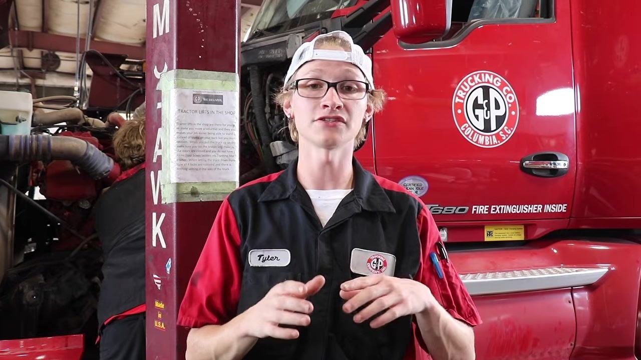 Technician standing in front of the truck, explaining the final inspection process. The truck and maintenance area are visible in the background.