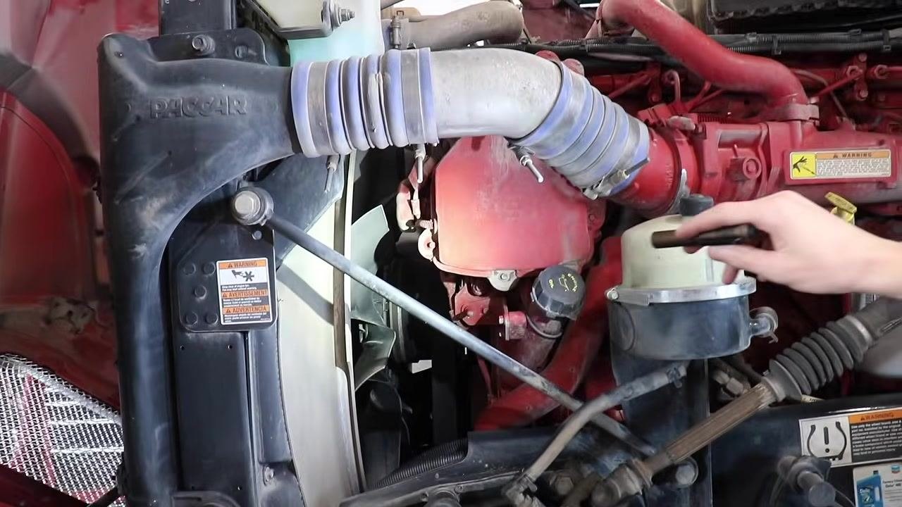 Mechanic's hand pointing at the radiator and power steering reservoir, showing hoses and radiator structure.