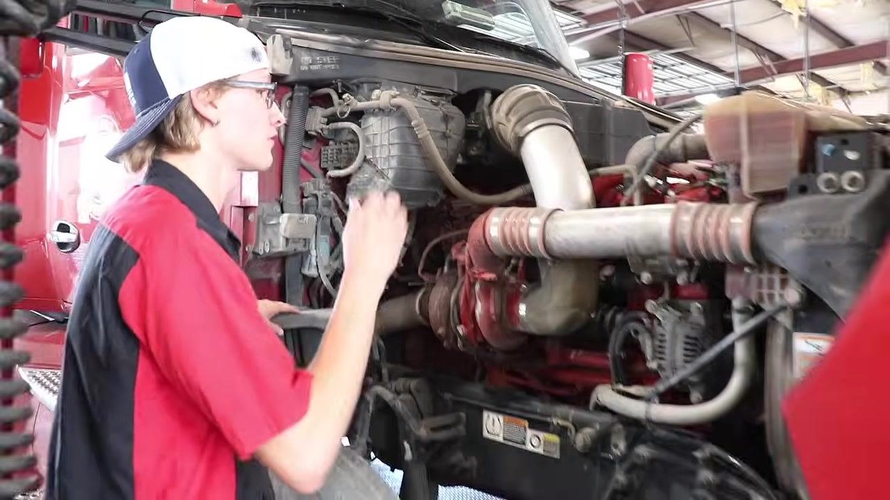 Mechanic leaning over the tire, inspecting brake pads and the brake chamber with a flashlight, with tire tread and air lines visible.