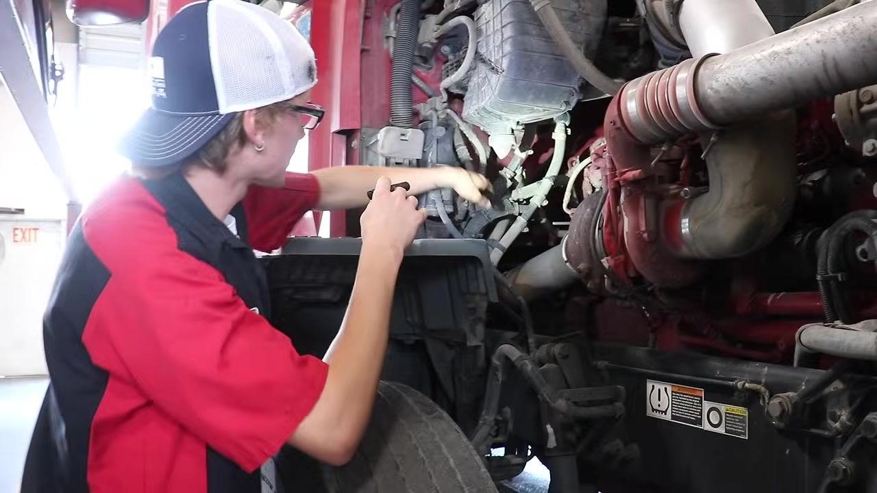 Mechanic using hands and a flashlight to inspect steering and brake lines, focusing on air lines and fittings.